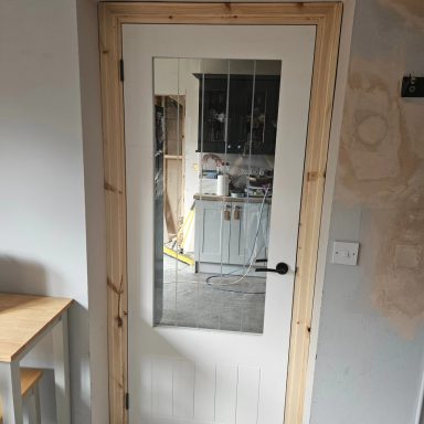 New door install with architrave White door with a glass panel framed in wood, leading to a room with furniture.