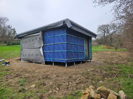 Small building with blue tarpaulin covering, set in a grassy area with trees nearby.